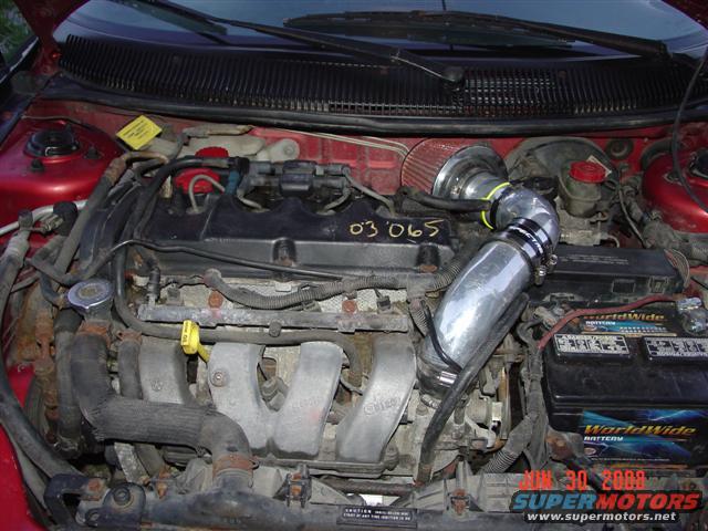 Engine bay of a red car, showing the engine block, battery, and an aftermarket cold air intake system
