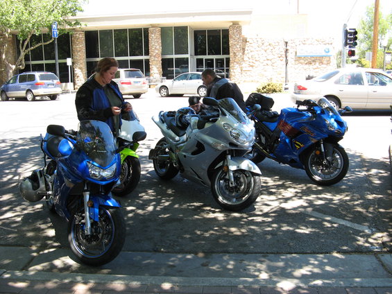 Three sport motorcycles parked on pavement, two blue and one silver. A woman stands near a blue motorcycle