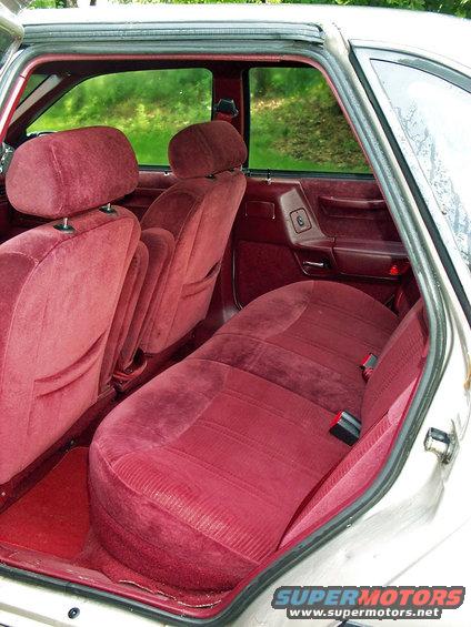 Rear interior of a car with red velour seats and door panels. Green trees visible through the windows