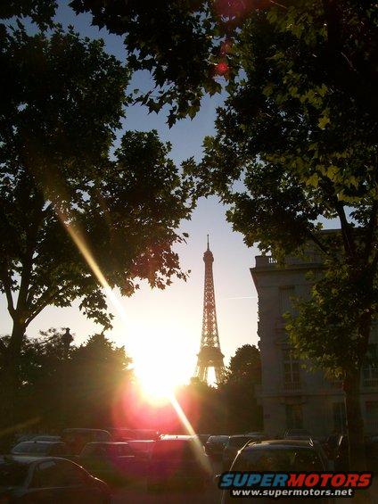 Eiffel Tower visible through trees at sunset, with a line of parked cars in the foreground