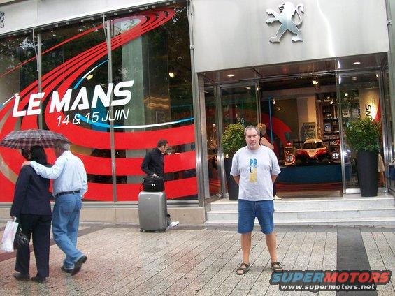 Man stands in front of a Peugeot dealership with a Le Mans advertisement in the window. People walk by with an umbrella