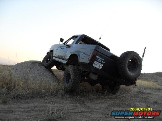 Gray 1990 Ford Bronco off-roading on a rock, rear-mounted spare tire visible