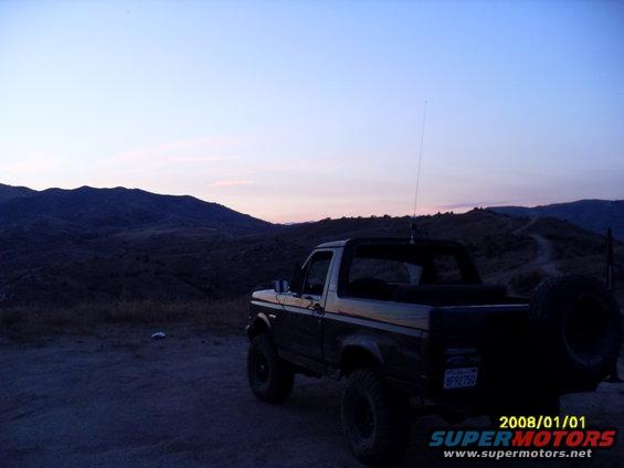 1990 Ford Bronco parked on dirt road, mountains in background at dusk. Spare tire mounted on rear