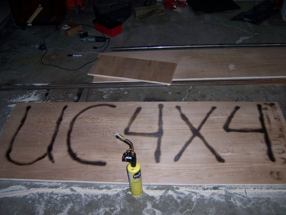 Plywood sheet with black spray-painted text "UC 4X4" and a yellow propane torch