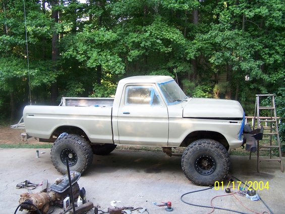 Tan 1977 Ford F-150 pickup truck with large tires, parked outdoors with trees in the background