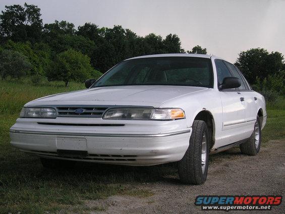 White Ford Crown Victoria sedan, front 3/4 view, parked on a dirt road with grass and trees in the background