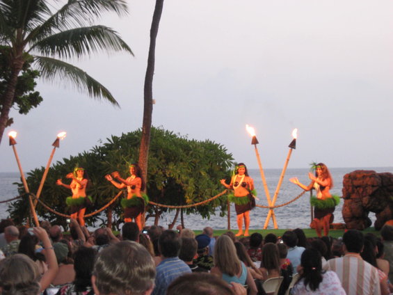 Four hula dancers perform on a stage by the ocean at dusk, holding flaming torches. An audience watches from below