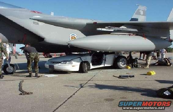 White car with open door positioned under a fighter jet wing