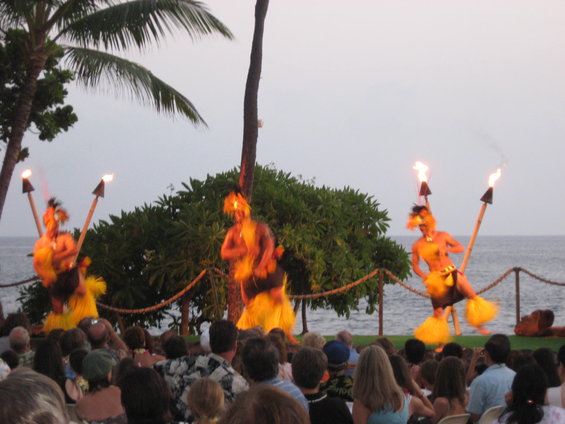 Luau performers with fire torches dance on a stage in front of an audience by the ocean
