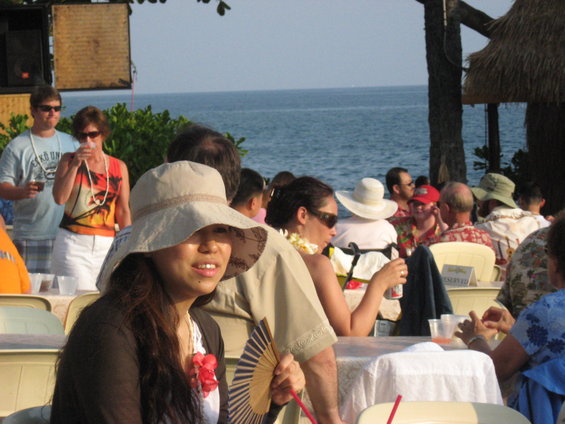 People seated at tables near the ocean, some wearing hats and leis