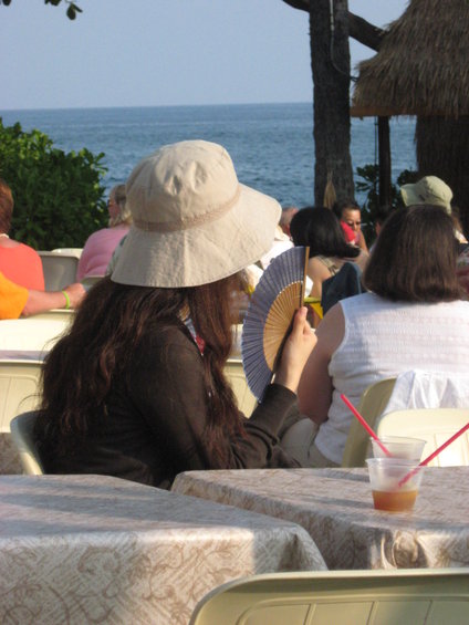People sit at tables near the ocean, one person fans herself