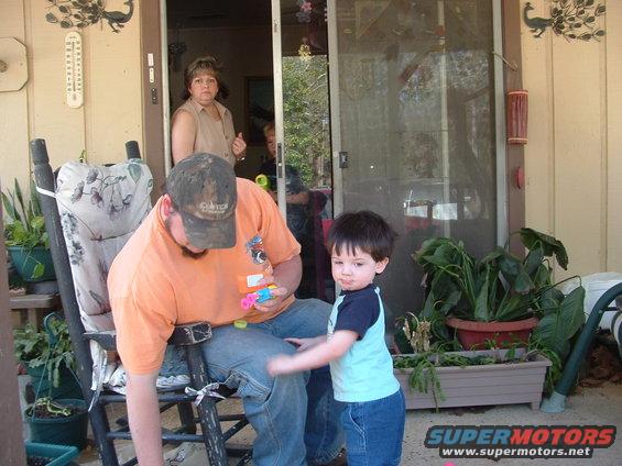 Man sitting in a rocking chair on a porch, holding toys, with a child standing nearby. A woman is visible in the doorway behind them