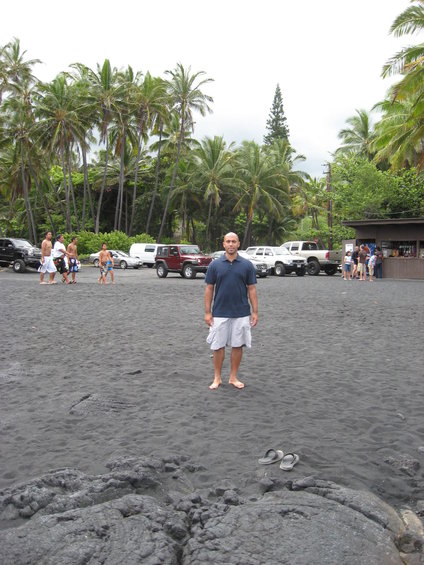 Man in blue shirt and white shorts stands on black sand beach, facing camera. Several parked cars and trucks are behind him