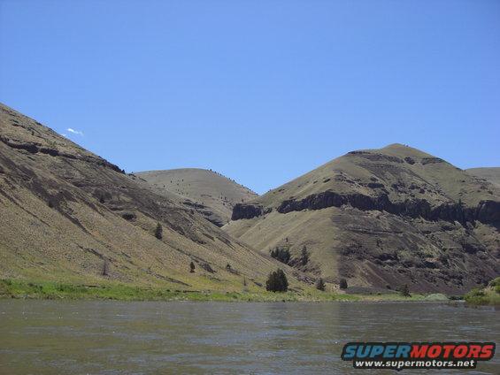 River flows past grassy banks with rolling, arid hills under a clear blue sky