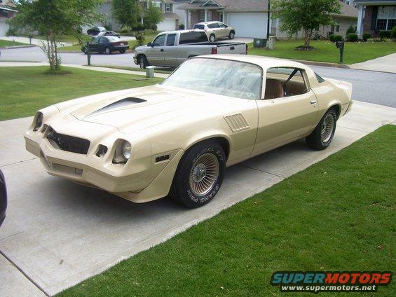 Tan coupe with a hood scoop parked on a driveway. It looks like a 1977 Chevrolet Camaro