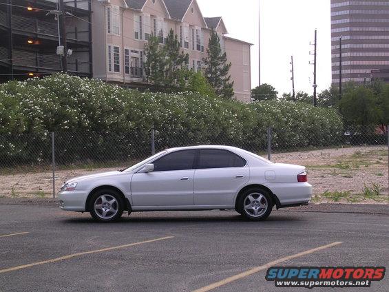 Side view of a white sedan parked in a lot. Buildings and trees are in the background
