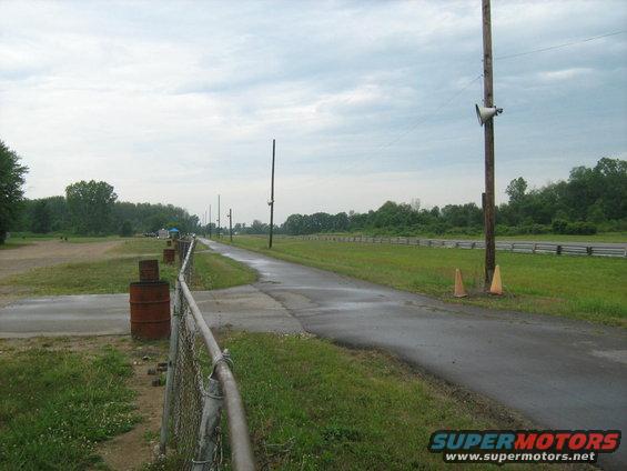 Drag strip with wet asphalt, grass, chain-link fence, barrels, and utility poles under a cloudy sky