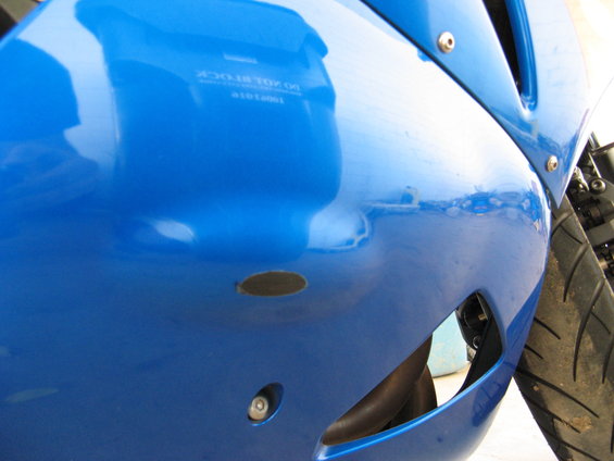 Blue motorcycle fairing with a scuff mark and a small hole. Reflection shows text