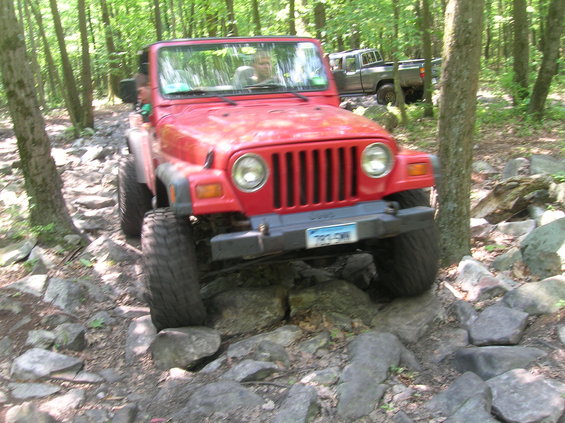 Red Jeep Wrangler driving over rocks on a wooded trail. A pickup truck is visible behind it