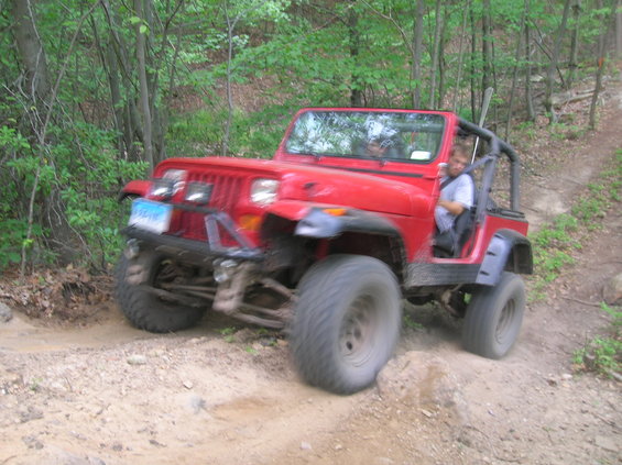 Red Jeep Wrangler off-roading on a dirt path, front-left tire angled up a rock