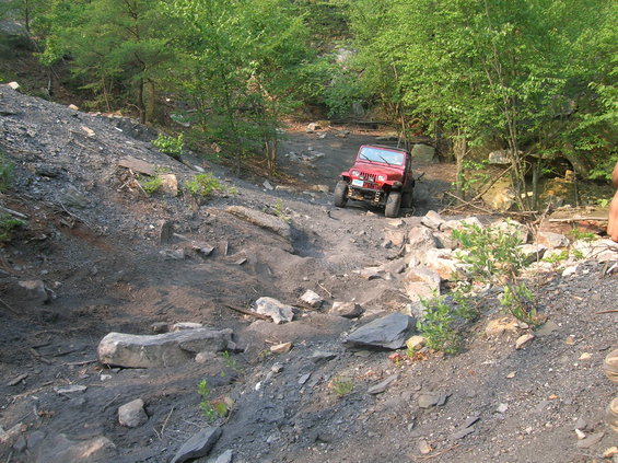 Red Jeep Wrangler navigating a steep, rocky off-road trail with trees on either side