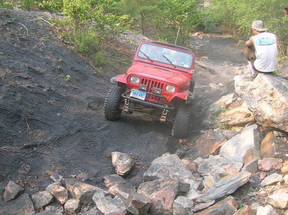 Red Jeep Wrangler navigating a rocky, off-road trail with a person sitting on a rock in the background