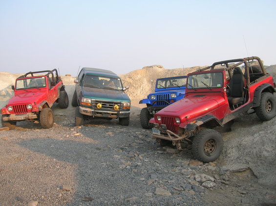 Four off-road vehicles, including red and blue Jeeps and a green SUV, parked on a rocky, uneven terrain under a clear sky