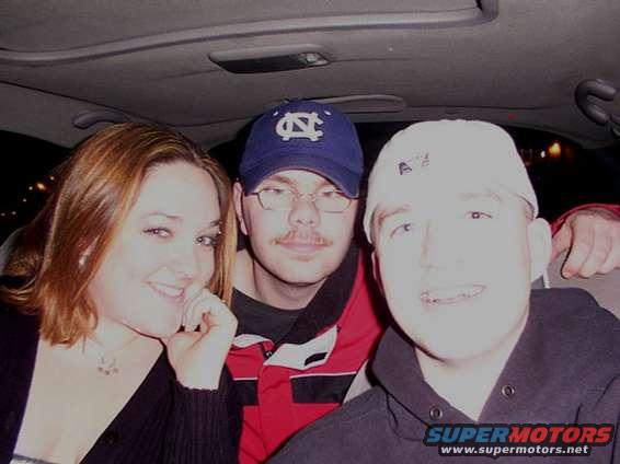 Three people inside a car, two men and a woman. One man wears a blue baseball cap