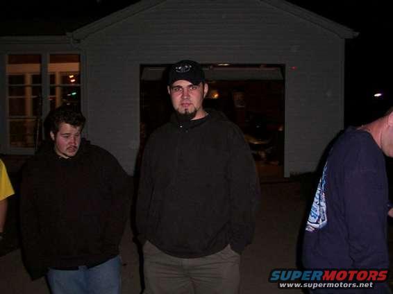 Three men stand in front of a garage at night. One man in the center wears a black baseball cap and goatee