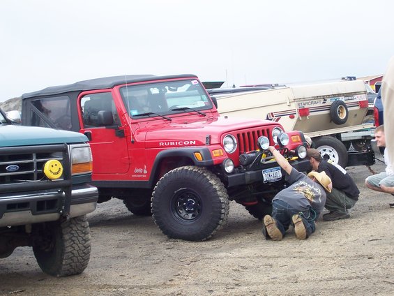 Red Jeep Wrangler Rubicon with aftermarket bumper and winch, people working on it. Green Ford Explorer in foreground