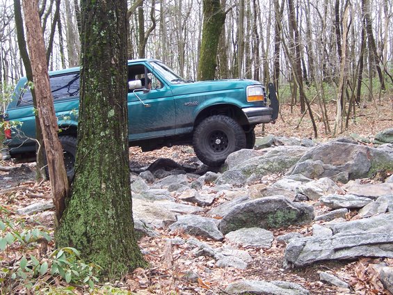 Green 1996 Ford Bronco navigating a rocky forest trail, front-right tire on a rock