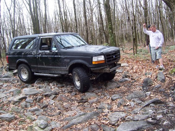 Black 1996 Ford Bronco driving over rocks in a wooded area. A person stands to the right