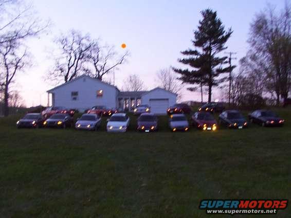 Multiple sedans parked in a row on a hill in front of a house at dusk
