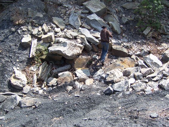 Person in brown hoodie and jeans stands on a rocky, coal-strewn slope