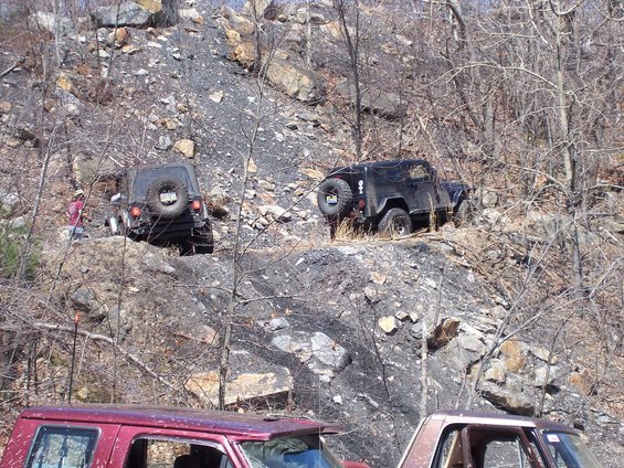 Two off-road vehicles are navigating a steep, rocky hill with sparse trees. A person stands near the left vehicle