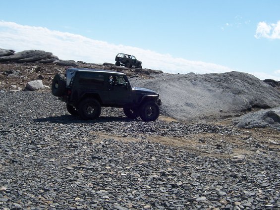 Two dark-colored off-road vehicles navigate a rocky, uneven landscape under a partly cloudy sky