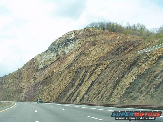 Highway passes by a large, layered rock formation under a cloudy sky