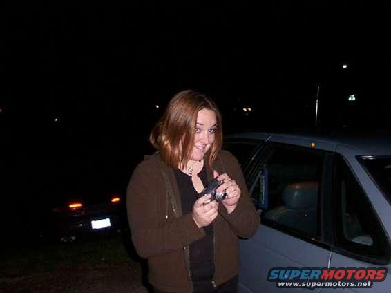 Woman holding camera next to a gray car, with another car in the background