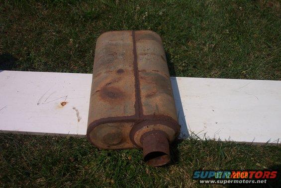 Rusty oval exhaust muffler with a single tailpipe, resting on a white board on grass
