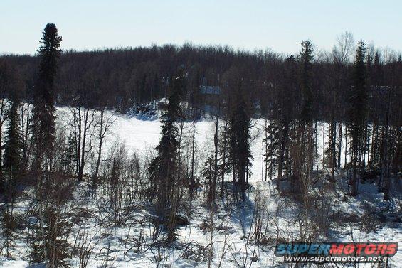 snowy landscape with bare trees and evergreens, a frozen lake or river in the background