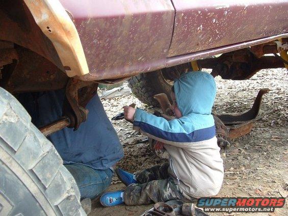 kid in blue hoodie helps someone work on the underside of a rusty vehicle