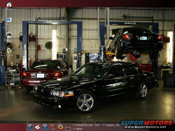 Black sedan in foreground, red sedan behind it, another black sedan on lift in background. Garage setting