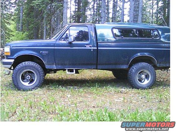 Dark blue pickup truck, side view, with a green camper shell, parked in a grassy area with trees in the background