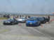 1985 Ford Mustang - Cars are staged on a race track, with cones marking the course and spectators in the background.