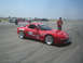 1985 Ford Mustang - A red Mazda RX-7 race car is parked on an asphalt track with other vehicles in the background.
