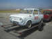 1985 Ford Mustang - A white Fiat 850 Coupe, numbered 903 and bearing 'Tire Rack' branding, is loaded onto a trailer, with a red pickup truck visible behind it.