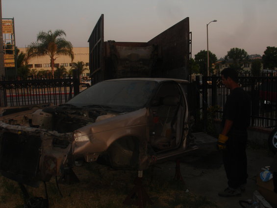 Damaged sedan on jack stands, front end removed, driver's door open. A man stands nearby
