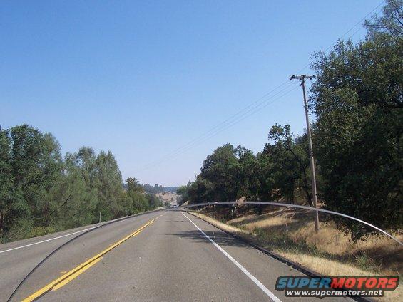 Driving down a two-lane highway with trees and hillsides on either side under a clear blue sky. A utility pole is visible on the right