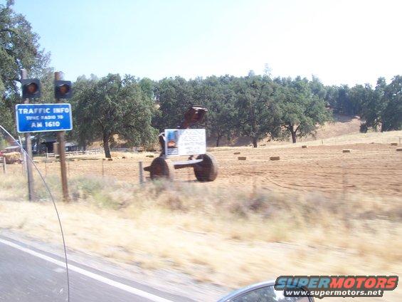Metal sculpture with large wheels and a sign in a field with hay bales and trees. Traffic info sign is visible