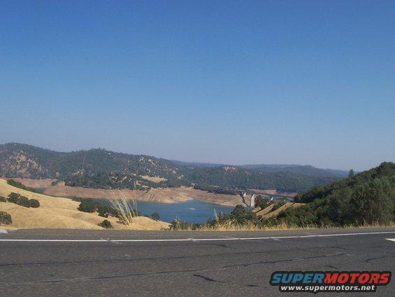 View of a lake and hills from a roadside, with a watermark 'SUPERMOTORS.NET'